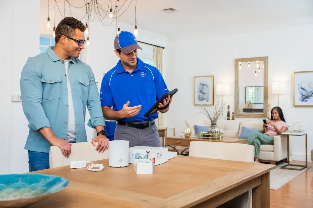 An ADT technician in a blue uniform uses a tablet to explain a home security system to a customer while standing near a table with equipment.